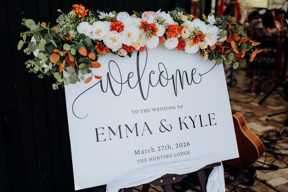 Wedding welcome sign for Emma & Kyle on March 27th, 2026 at The Hunting Lodge, adorned with white and orange floral arrangement.