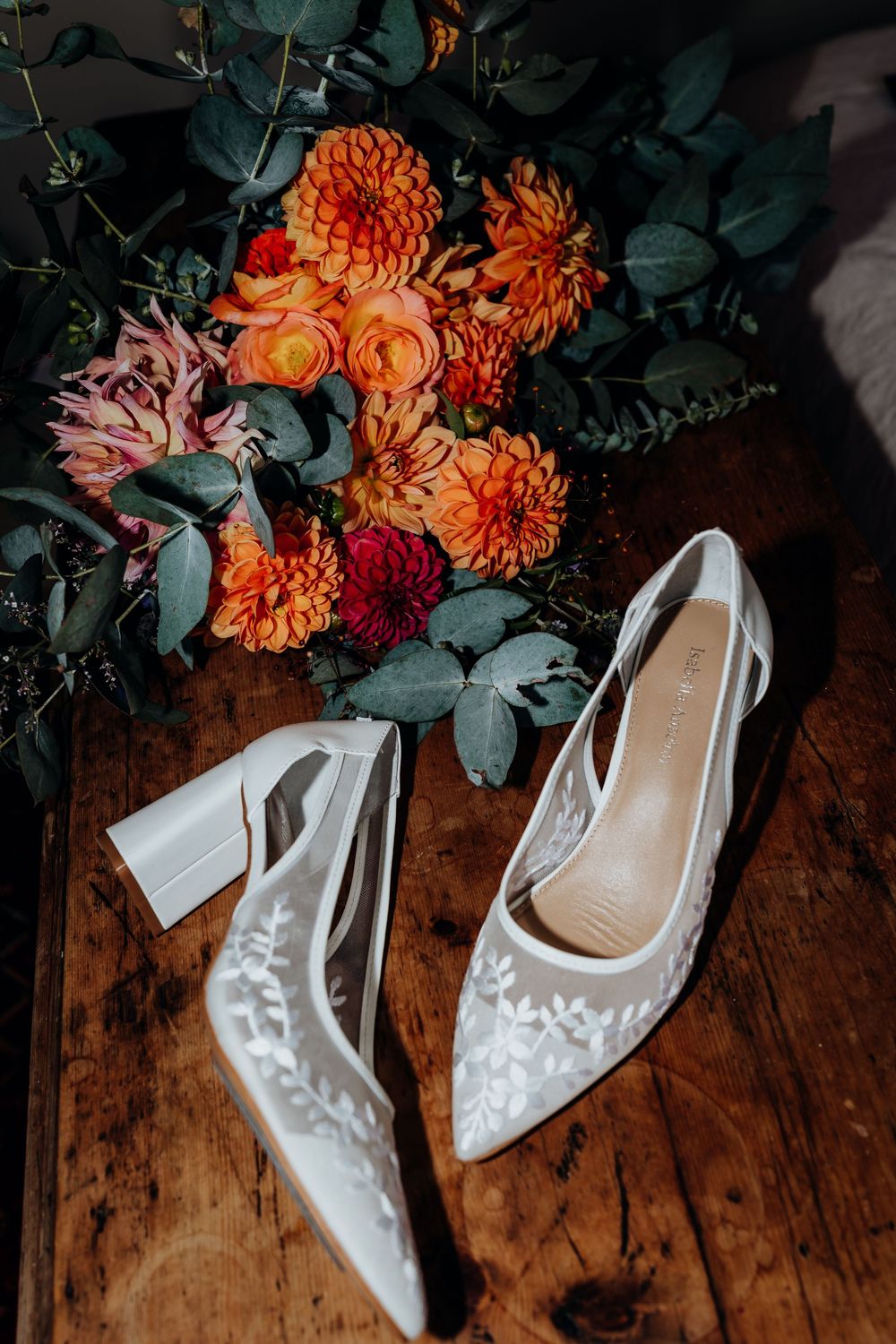 White lace bridal heels beside a vibrant orange and red wedding bouquet on a rustic wooden surface.