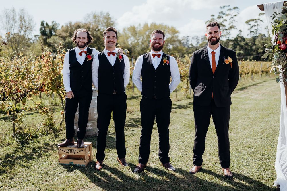 Four groomsmen in black suits with bow ties and boutonnieres standing outdoors at a vineyard wedding.