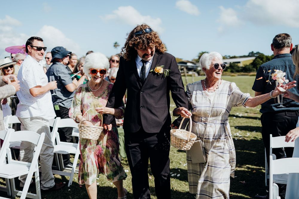 Groom walking down outdoor wedding aisle with two elderly women flower girls in sunny ceremony.