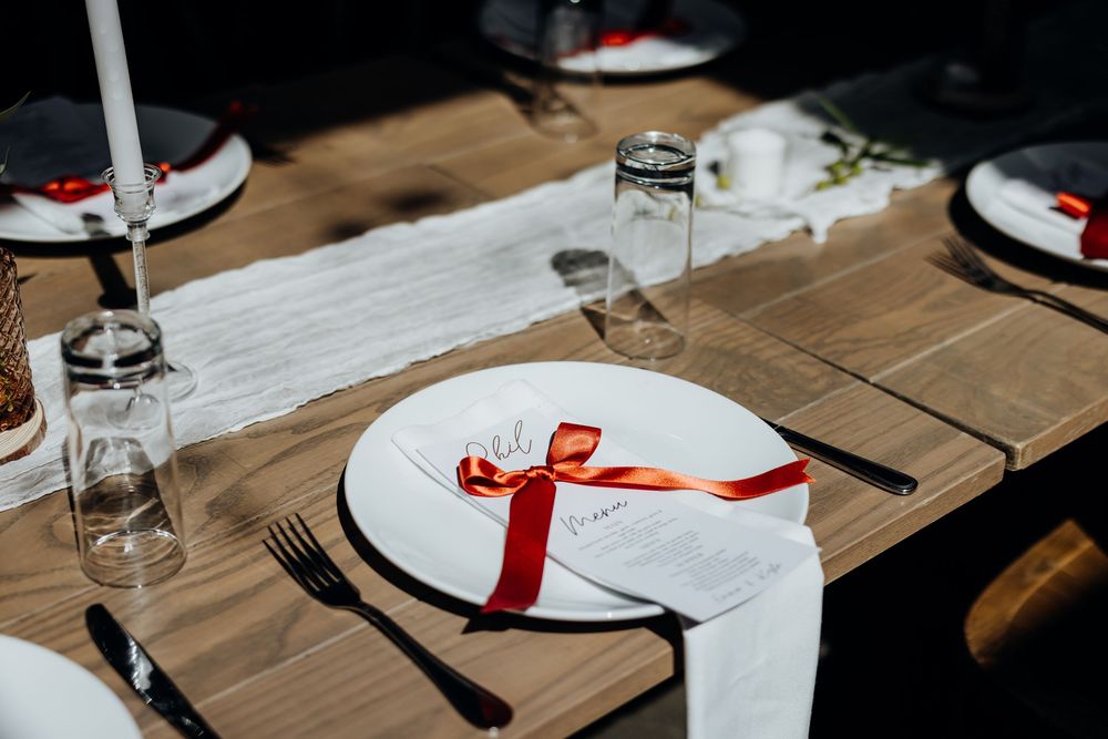 Elegant wedding reception place setting with white plate, red ribbon-tied menu card, and candle on rustic wooden table.