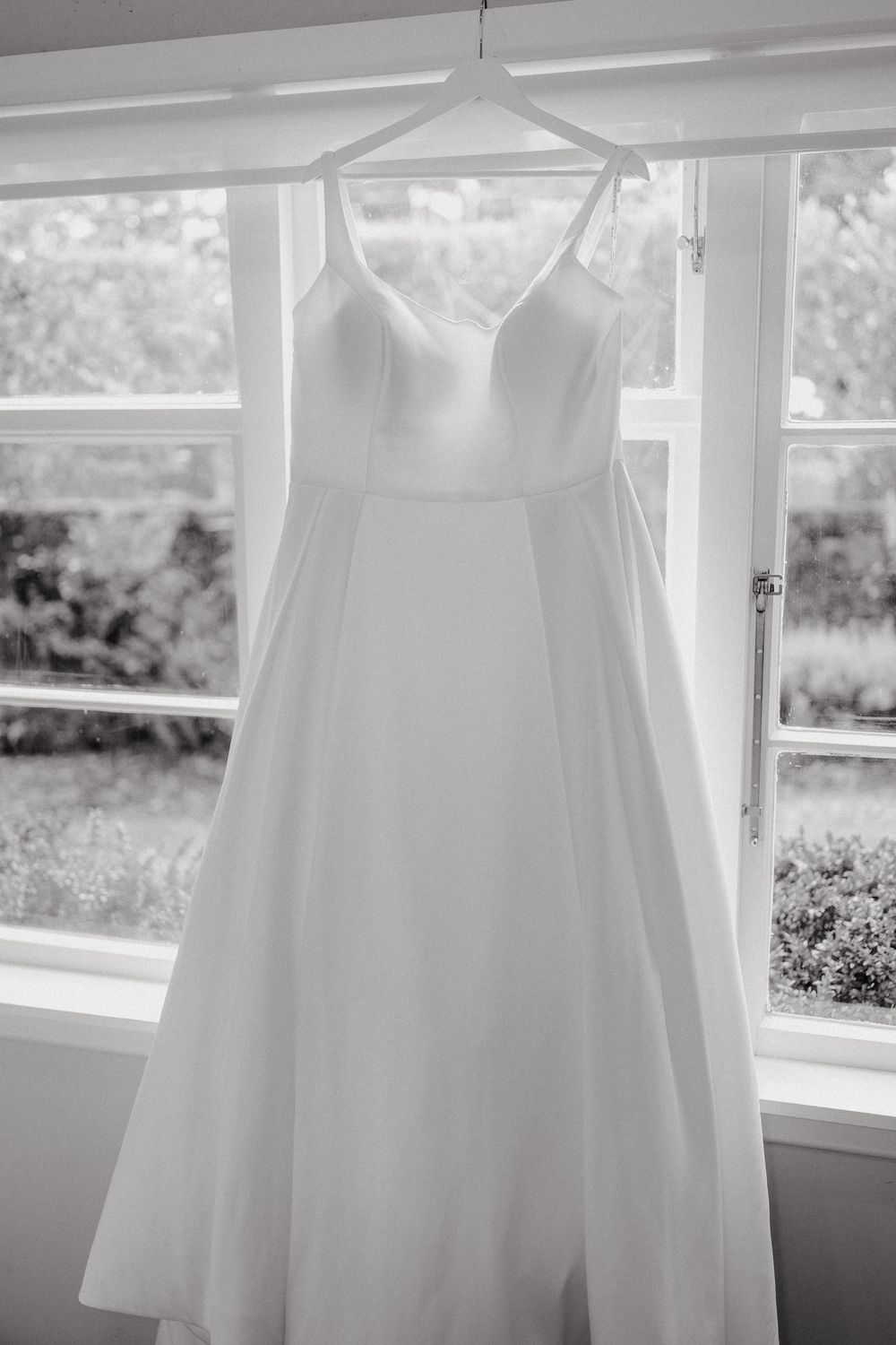 Elegant white wedding dress with spaghetti straps hanging on a hanger in front of a bright window.