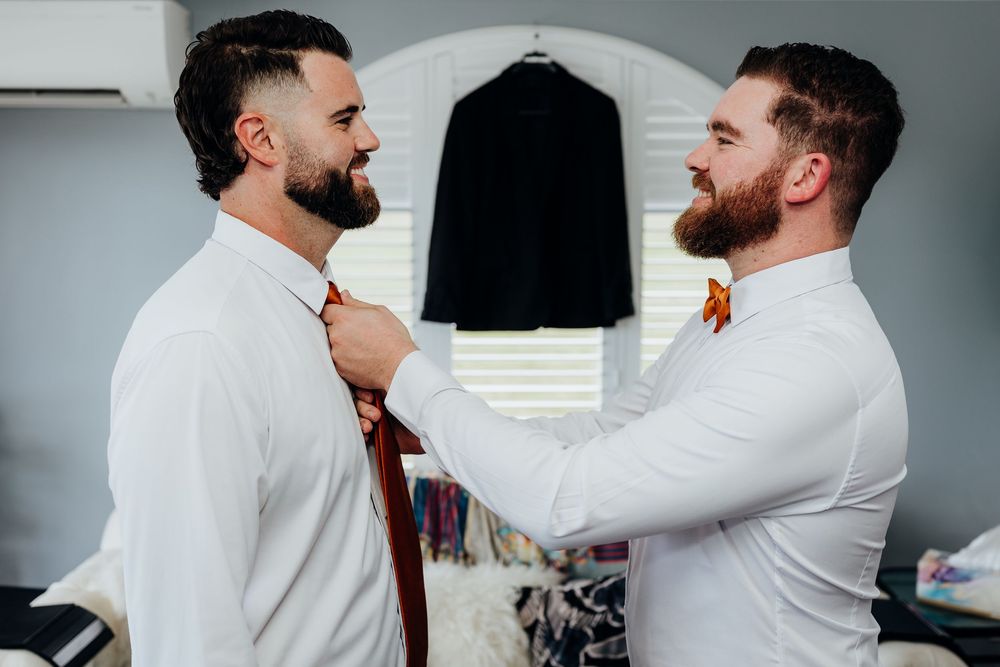 Two bearded groomsmen in white shirts help each other with a tie while preparing for a wedding ceremony.
