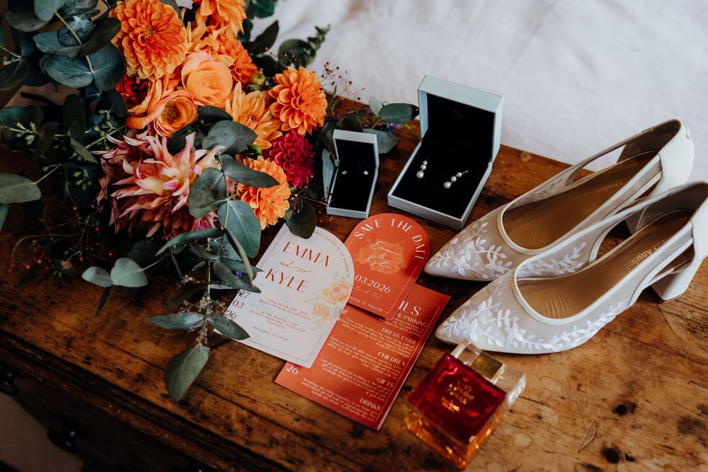 Wedding flat lay featuring orange floral bouquet, lace heels, jewelry boxes, and orange invitation suite on rustic wood.