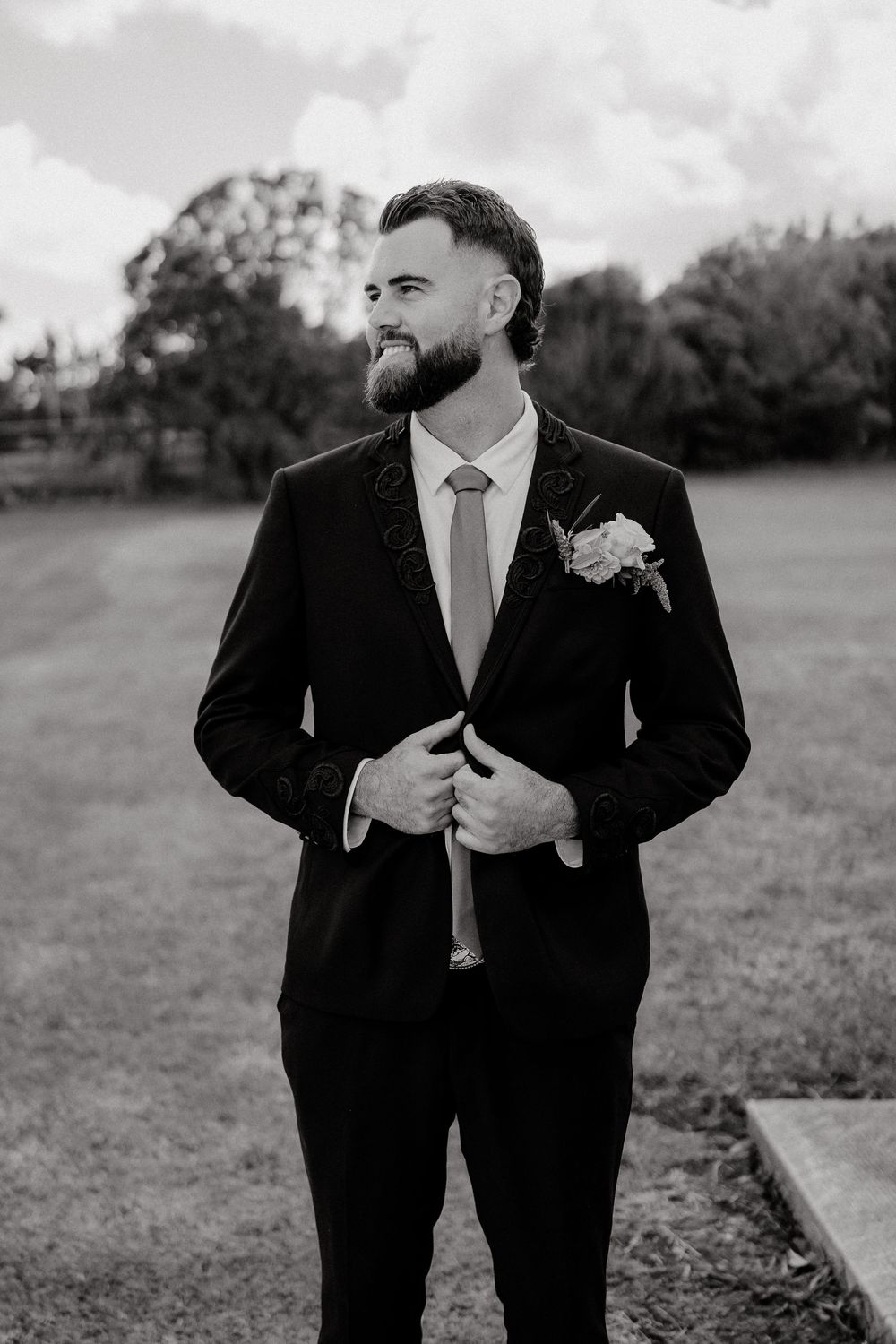 Bearded groom in black suit with boutonniere adjusting jacket outdoors in black and white photo.