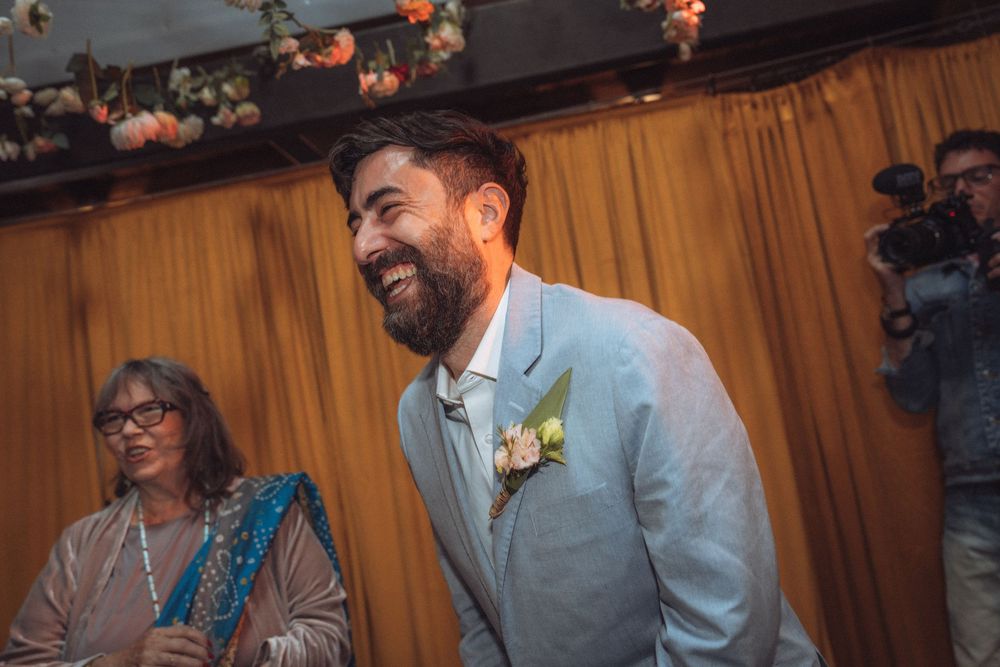 Smiling groom in light blue suit with boutonniere laughing joyfully at wedding ceremony indoors.