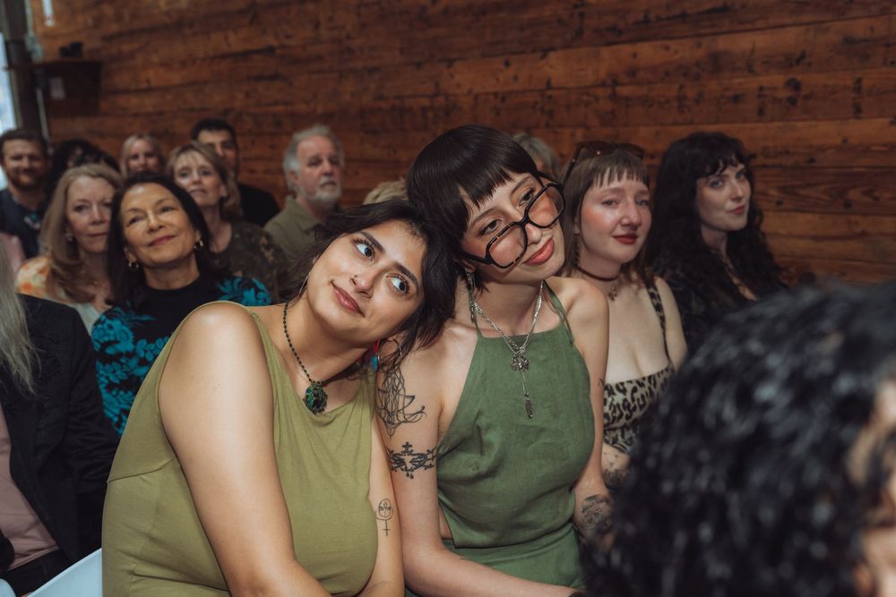 Group of smiling wedding guests in green outfits watching ceremony inside rustic wood-paneled venue.