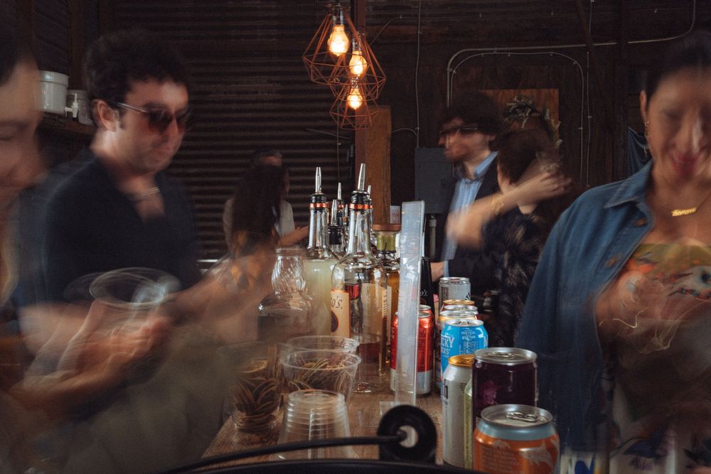 Busy bar scene with blurred motion of patrons and bartender surrounded by bottles and drinks in a dimly lit venue.