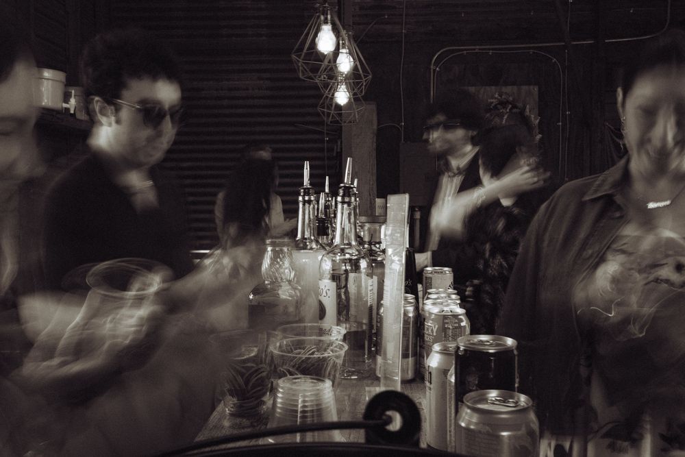Blurry black and white photo of people gathered at a dimly lit bar with bottles and glassware on the counter.