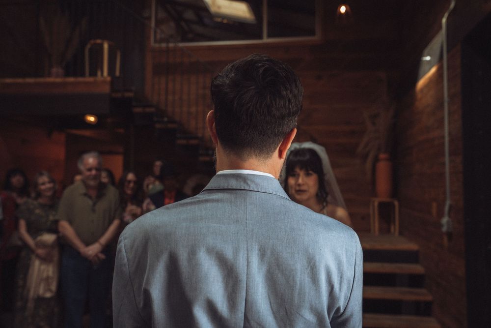 Groom stands at the altar facing his bride in a rustic wooden wedding venue with guests watching.