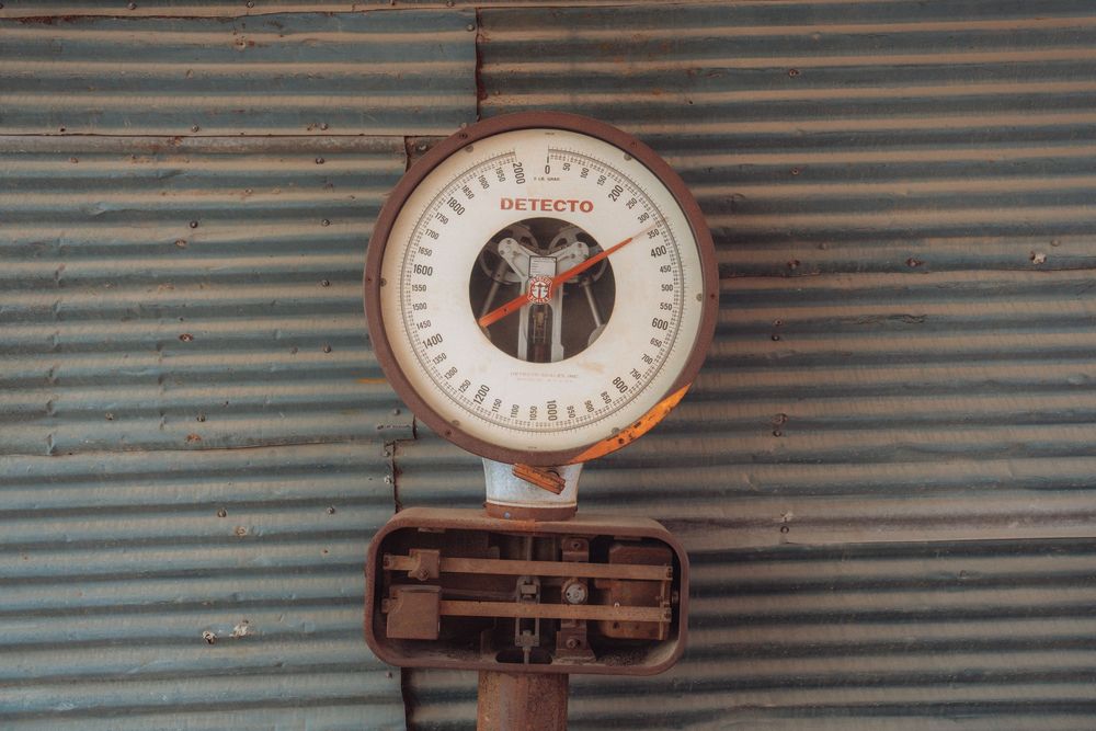 Vintage Detecto hanging scale with round dial face mounted on corrugated metal wall, showing signs of rust and age.