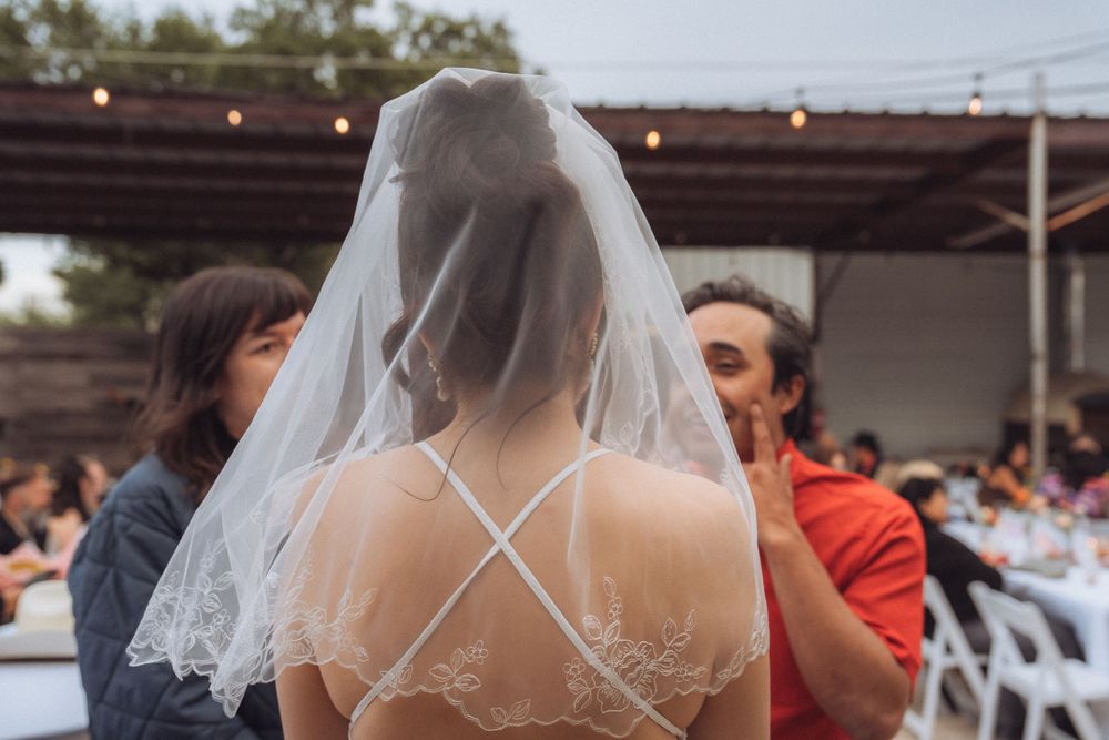 Bride in lace backless wedding dress with veil at outdoor reception, guests seated at tables in background.