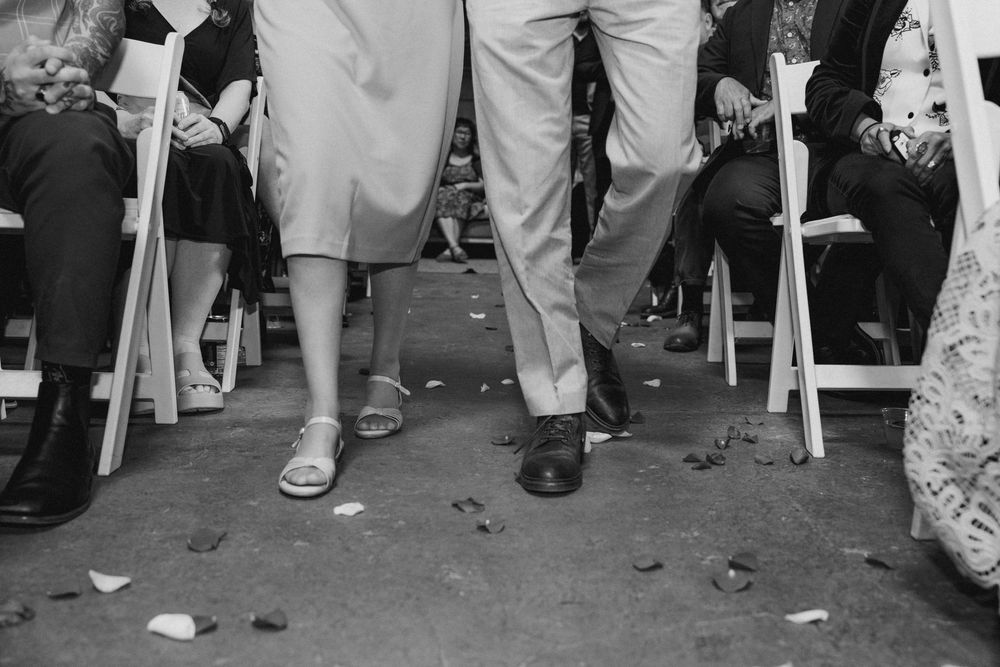 Black and white photo of a couple walking down the aisle at a wedding ceremony, rose petals on the ground.