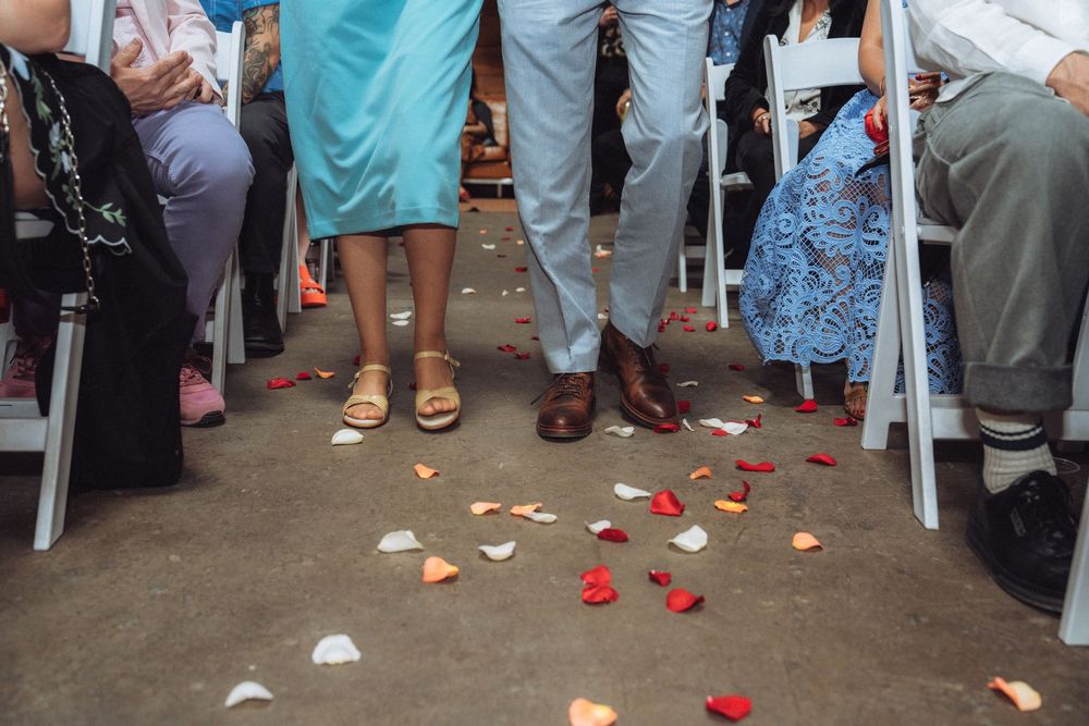 Wedding ceremony aisle with rose petals scattered on ground, guests seated on white chairs, couple standing together.