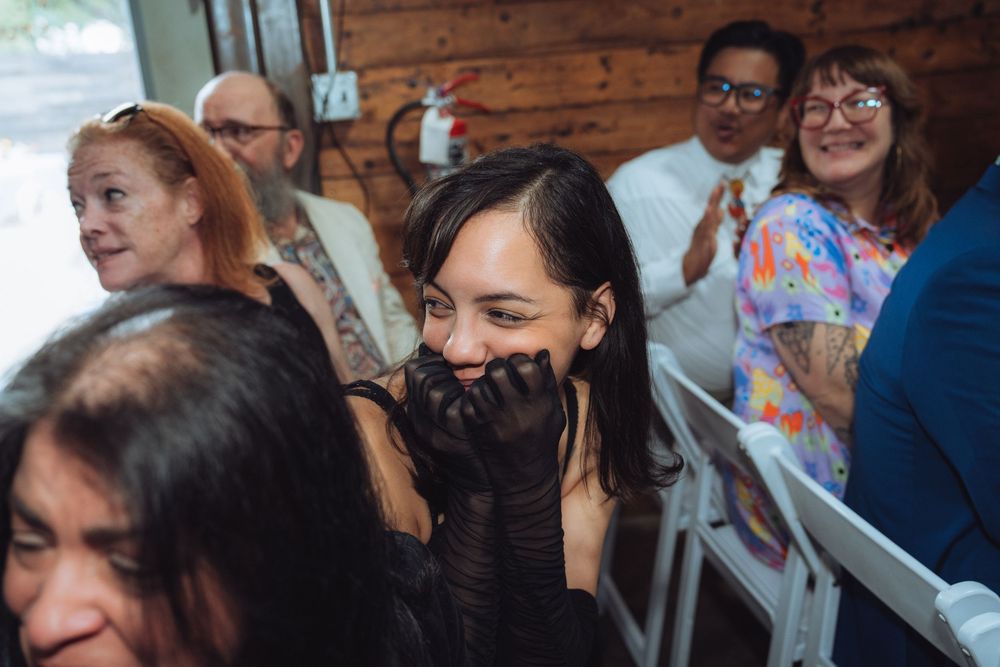 Guests laughing and smiling at a casual indoor event, woman in black covering mouth with gloved hands.