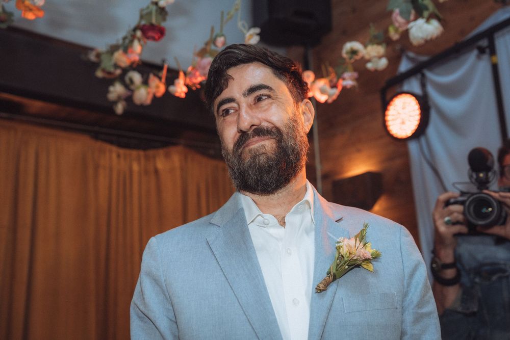 Smiling groom with beard wearing light gray suit and floral boutonniere at indoor wedding ceremony.