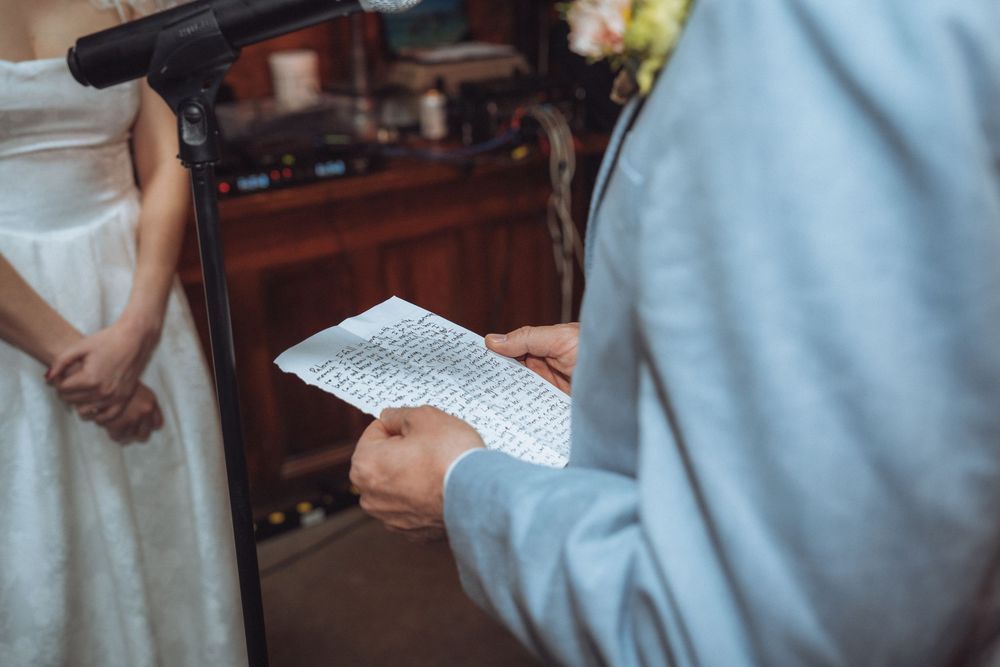 Groom reading handwritten wedding vows from paper during ceremony, bride standing beside him in white dress.
