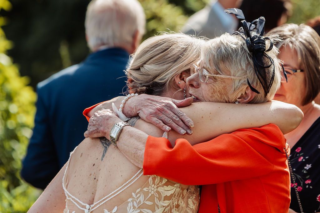 Two elderly women share a warm embrace at an outdoor wedding celebration, one wearing red jacket.