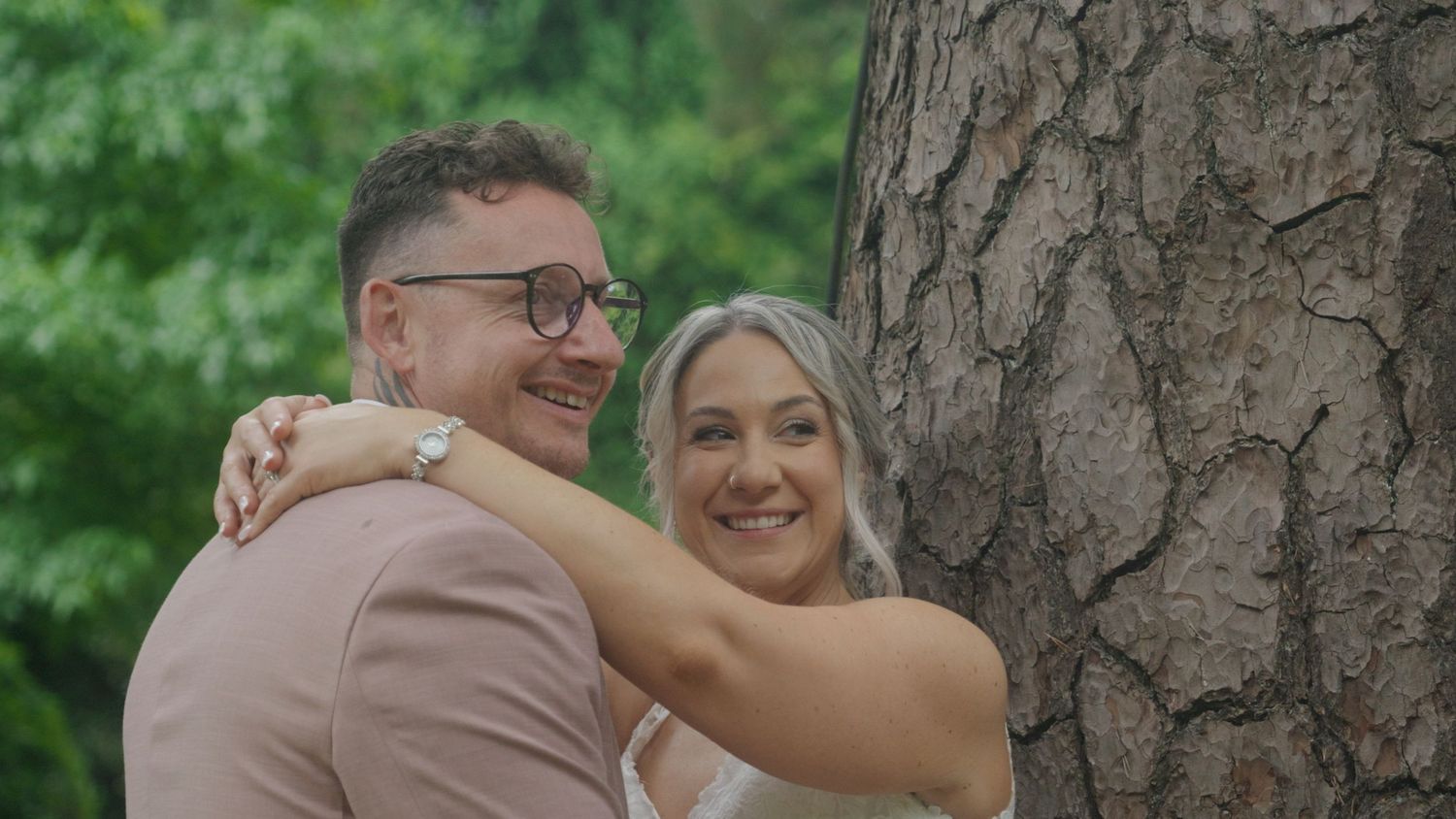 Couple embracing and smiling by a tree in a lush green park setting.
