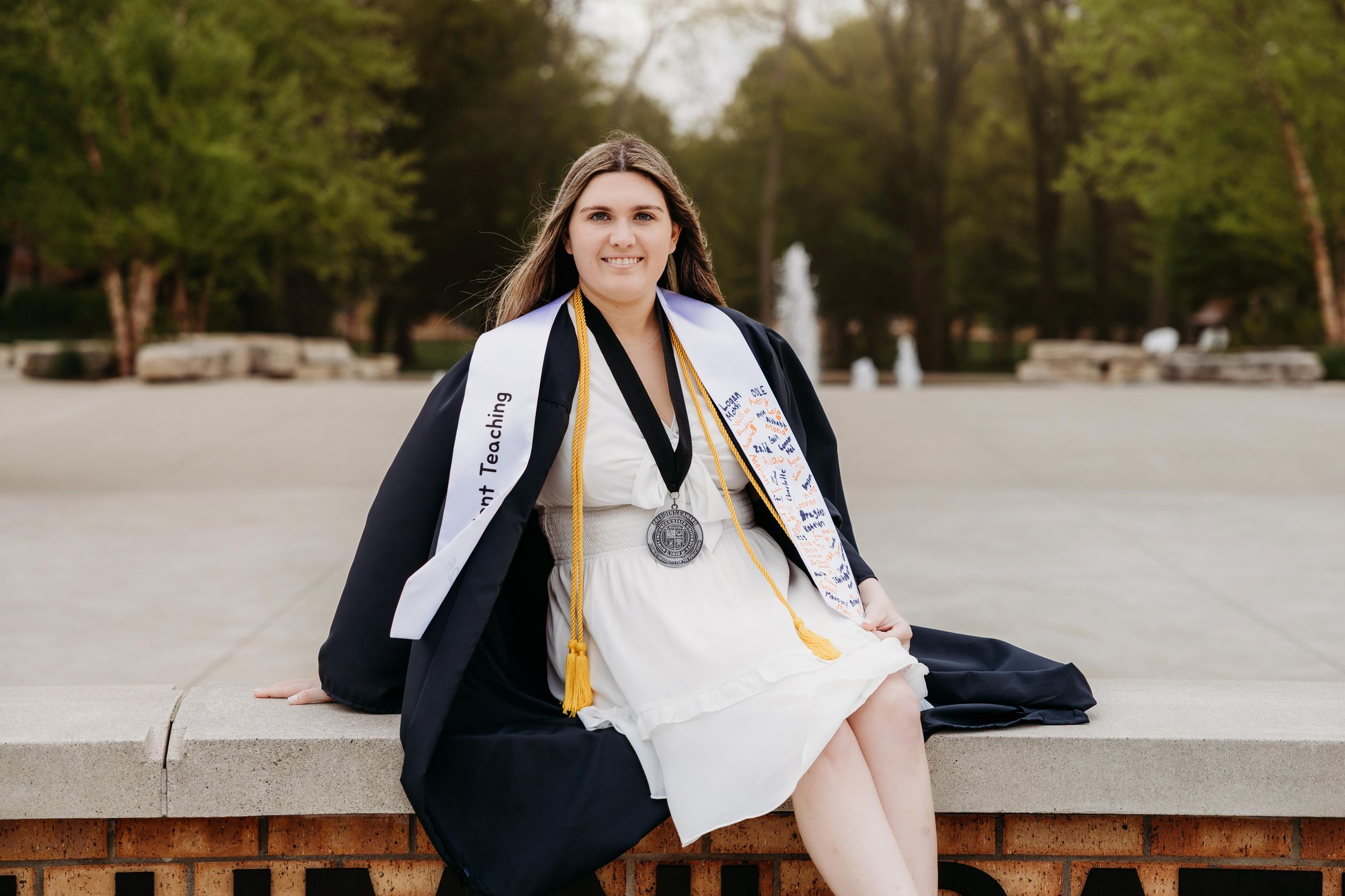 College graduate smiling in black gown, white stole, and medal, seated outdoors near a fountain.