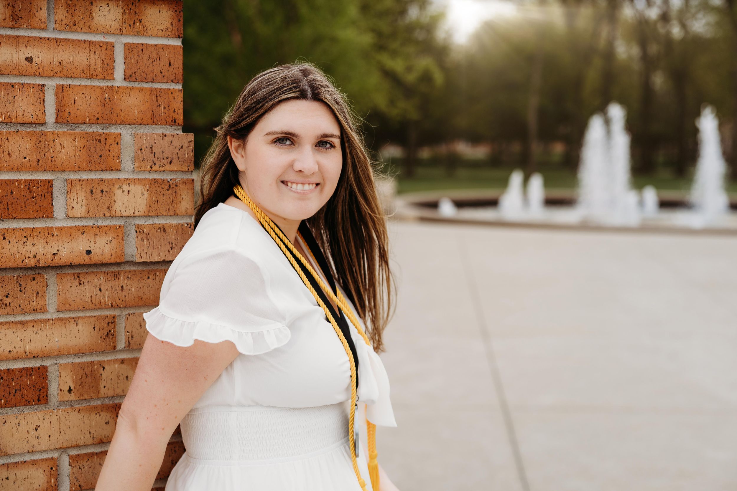 Young woman in white dress with gold graduation cords smiling outdoors near brick wall and fountain.