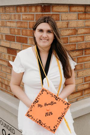 Smiling graduate holding orange cap reading Ms. Hamilton 2026, wearing white dress with gold and black honor cords.
