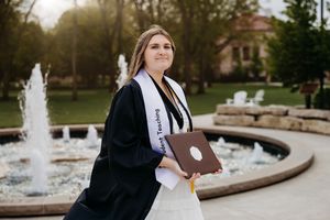 College graduate in black gown and white Teaching stole holds diploma by fountain on campus.