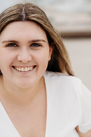 Smiling young woman with brown hair wearing a white top, portrait photo outdoors.