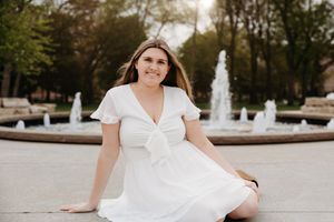 Young woman in white dress sitting by a decorative fountain outdoors, smiling warmly on a sunny day.