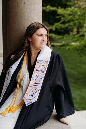 Young woman in black graduation gown and decorated stole sits against a stone column outdoors.