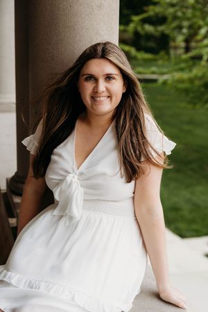 Young woman in white dress smiling outdoors near stone column with green garden background.