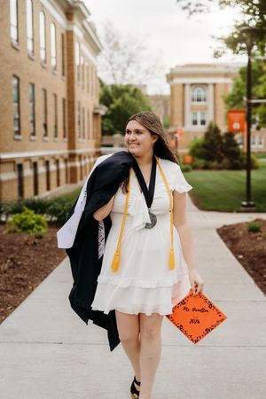 A smiling graduate in a white dress walks on campus, holding an orange cap and draping her black gown over her shoulder.