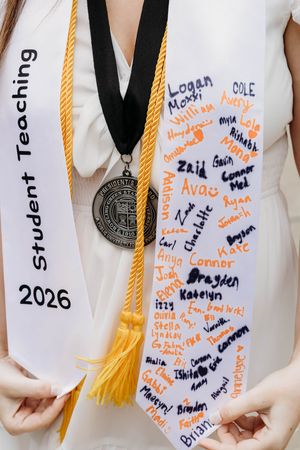 Graduate displaying Student Teaching 2026 sash with student signatures and gold honor cord with medallion.
