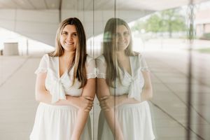 Young woman in white dress smiling and leaning against a glass wall with her reflection visible beside her.