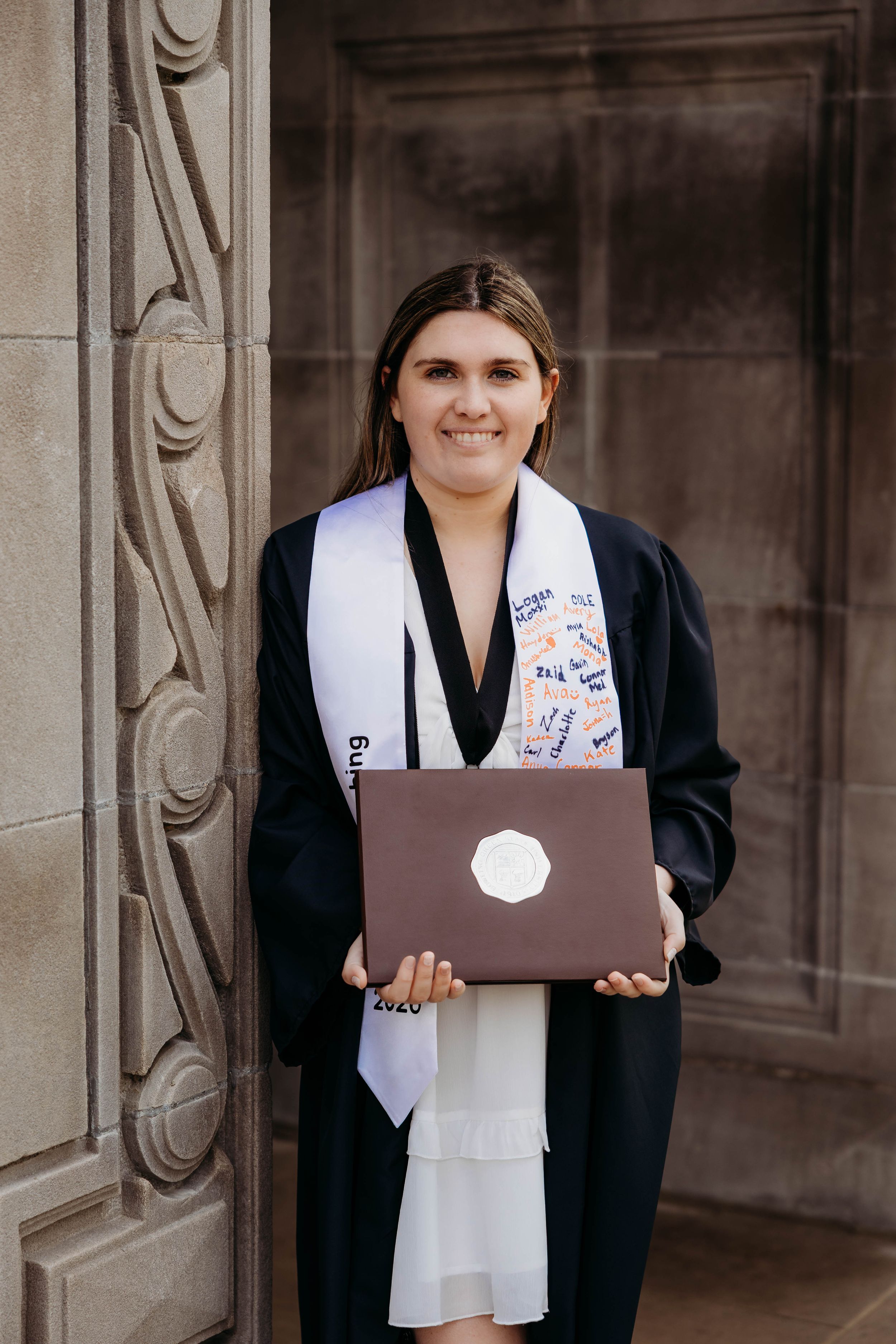 Smiling graduate in black gown holds diploma wearing white stole with patches outside stone building.