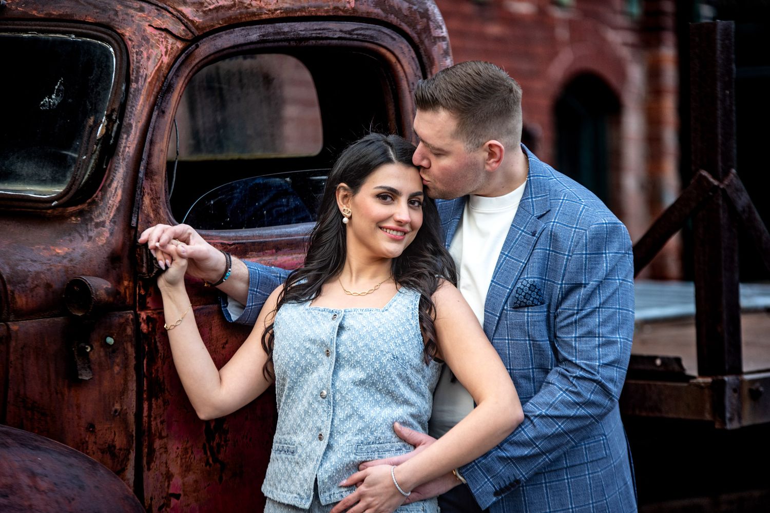 Couple posing by a rustic vintage truck, woman in grey outfit smiles while man in blue suit kisses her cheek.