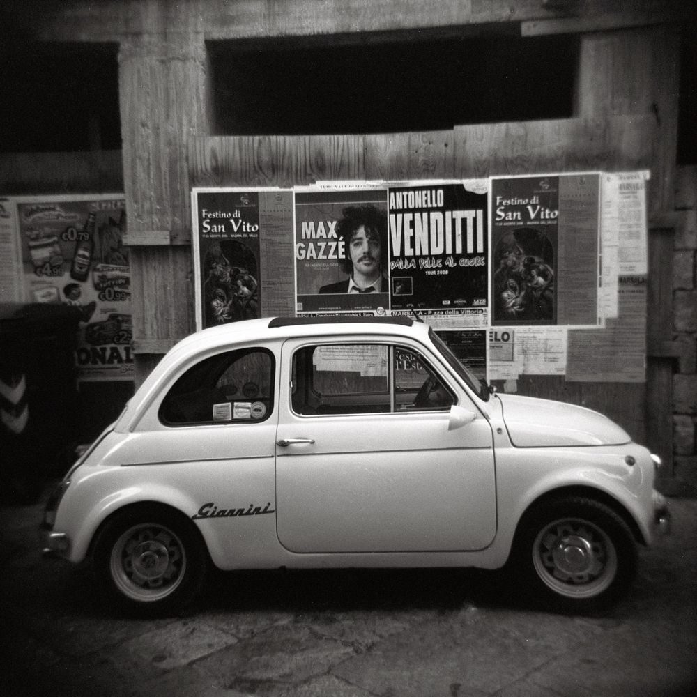 Vintage Fiat 500 parked in front of Italian concert posters in black and white photography.