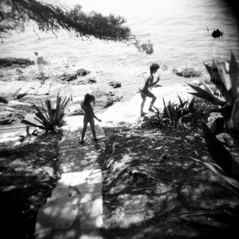 Black and white photo of children playing on rocky shoreline surrounded by agave plants and calm sea.
