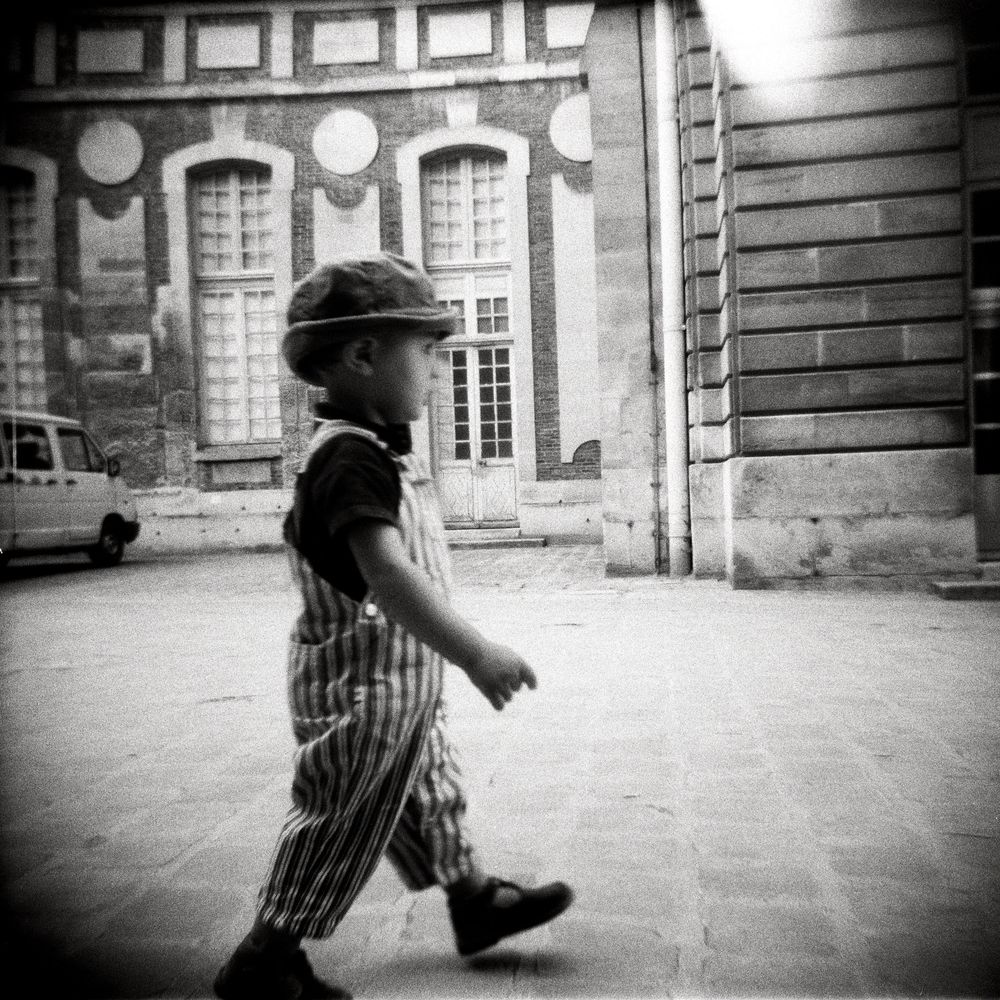 Black and white photo of a young child in striped overalls and hat walking across a cobblestone plaza near a building.