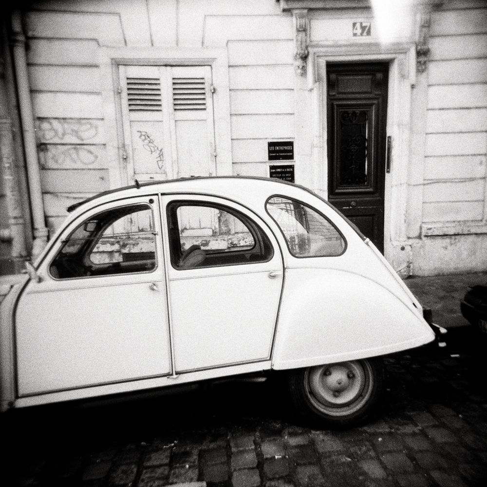 Black and white photo of a vintage Citroen 2CV parked on cobblestone street in front of classic European building.