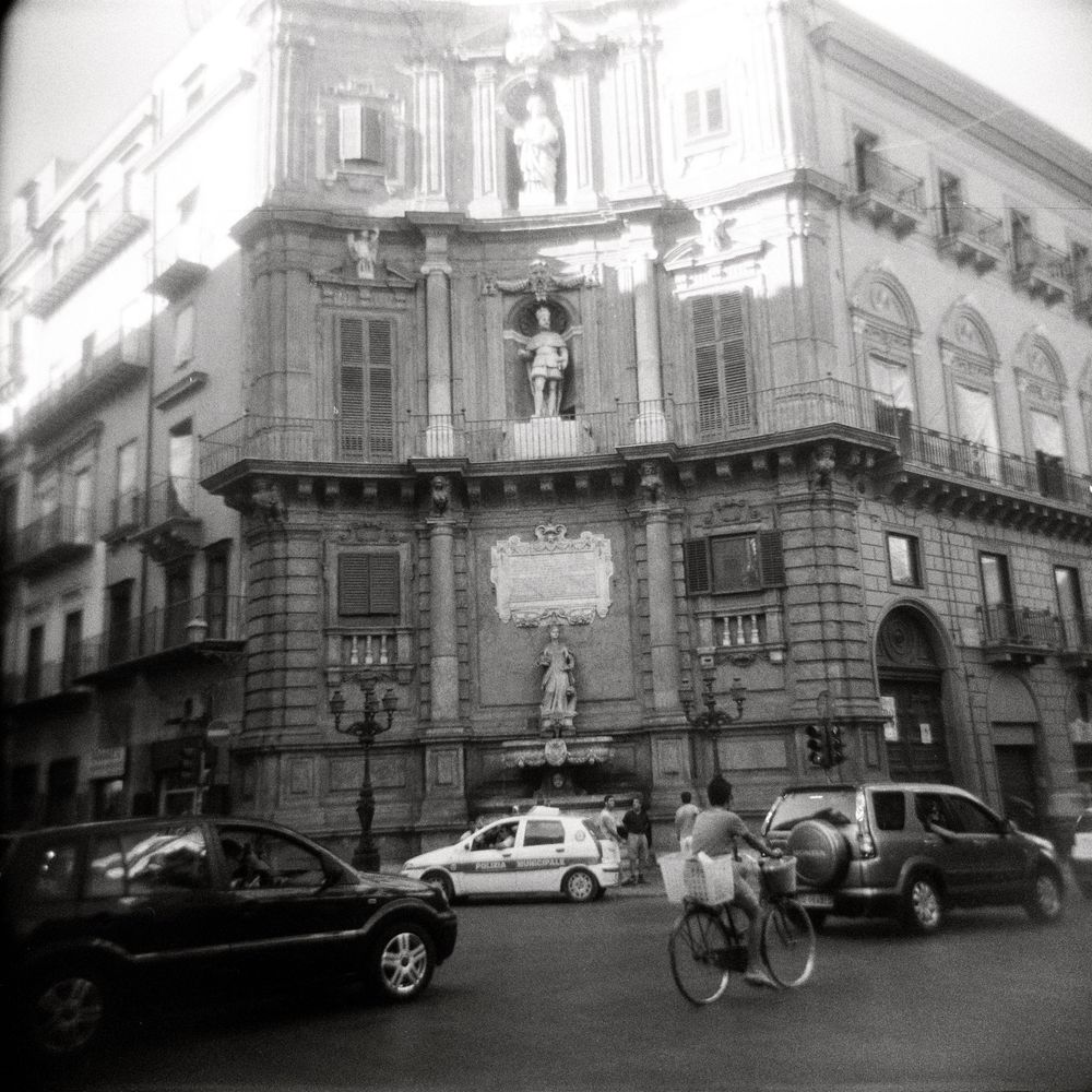 Black and white photo of a historic baroque building in Palermo, Italy, with cars and a cyclist in the foreground.