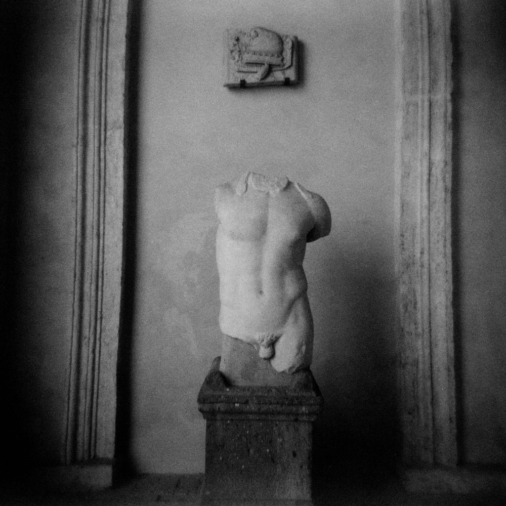 Black and white photo of a headless ancient Greek marble torso sculpture on a pedestal in a museum.
