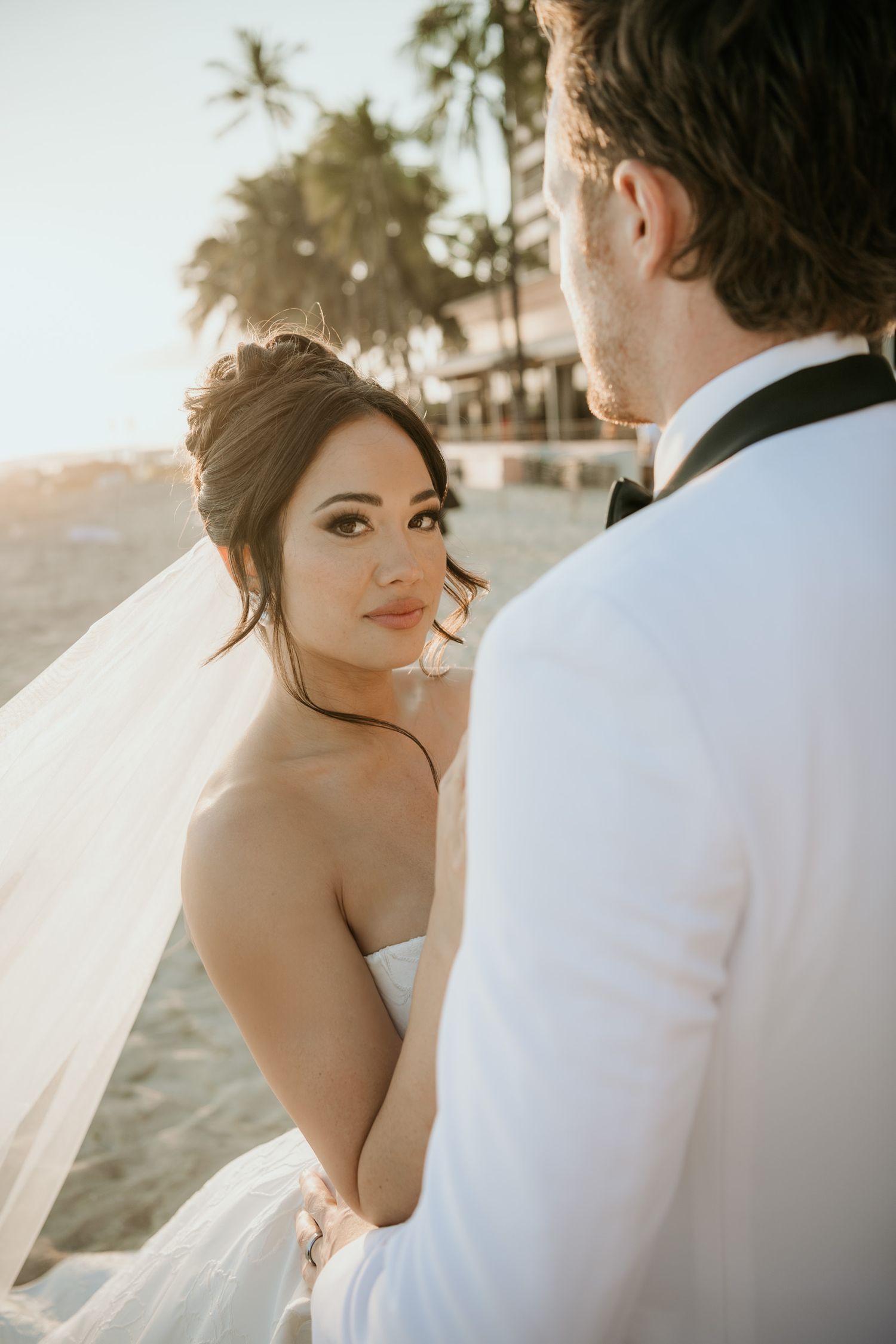 Bride in strapless gown and veil gazes over shoulder at groom in white suit during golden hour beach wedding.