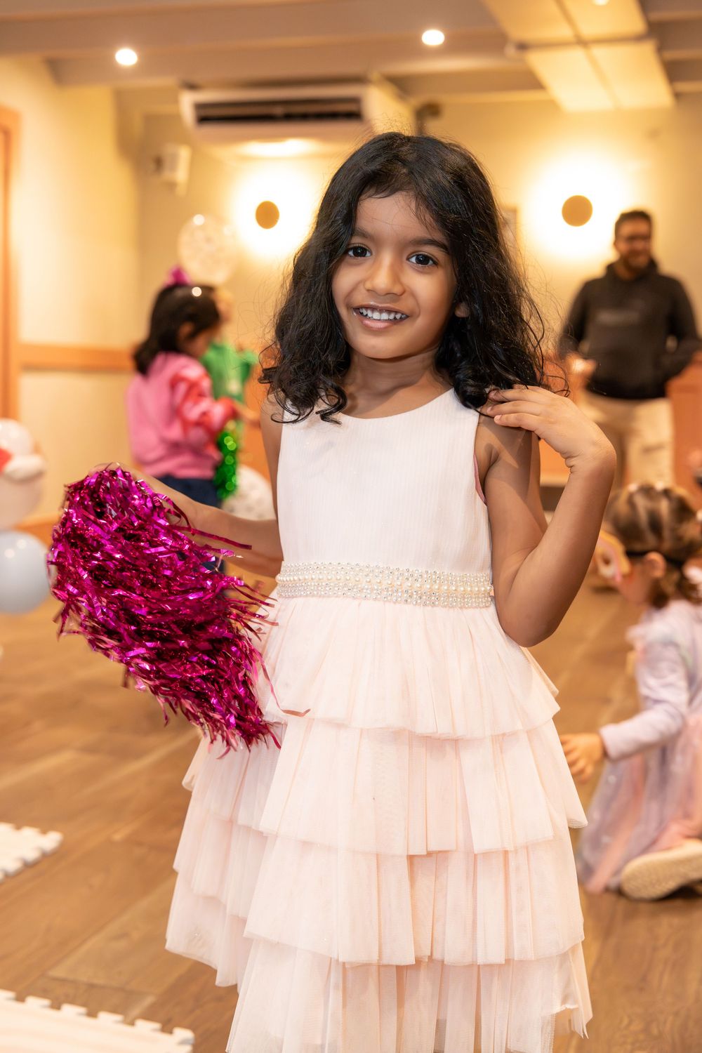 Young girl in white ruffled dress holding pink pom-pom at a birthday party, smiling joyfully.