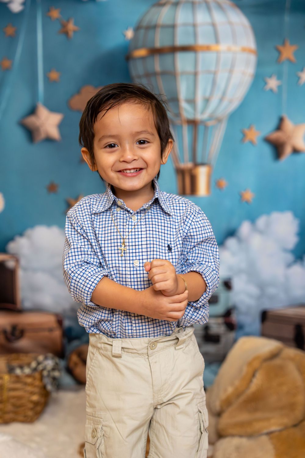 Smiling young boy in blue plaid shirt and khaki pants poses before a whimsical hot air balloon backdrop.