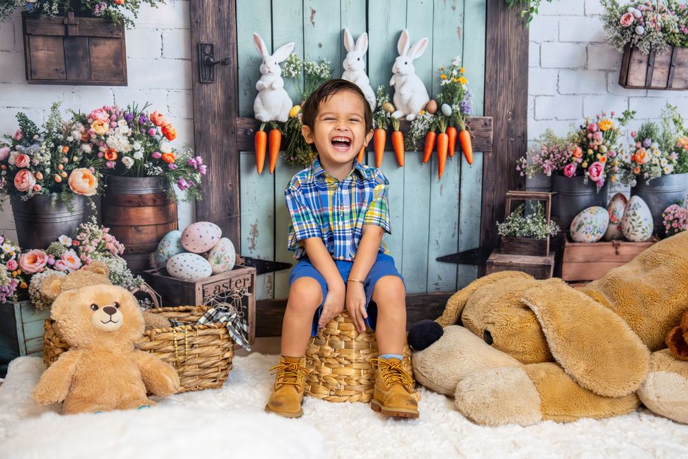 Laughing boy in bunny ears sits among Easter decorations, stuffed animals, and colorful flowers in a festive spring photo.