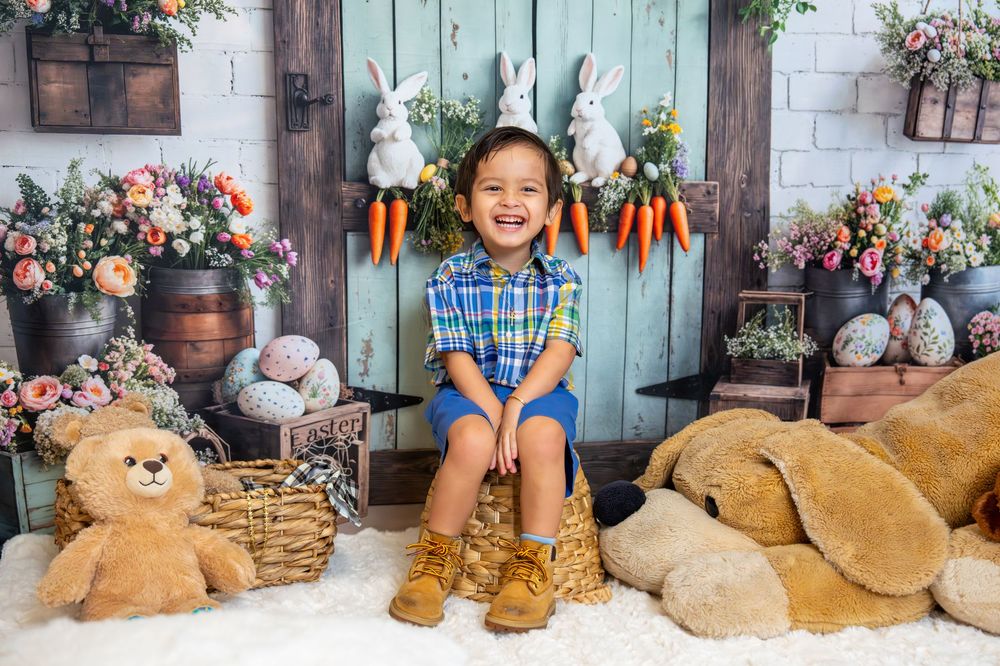 A joyful young boy in a plaid shirt sits smiling among Easter decorations, stuffed bunnies, and colorful spring flowers.