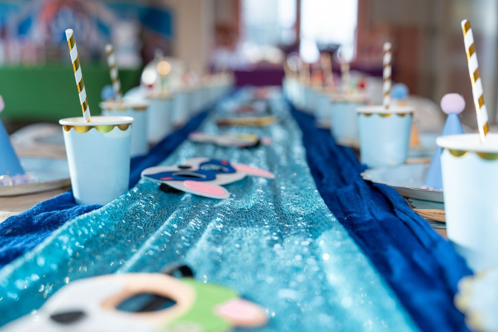 Festive party table with blue sequin runner, paper cups with striped straws, and colorful decorations.