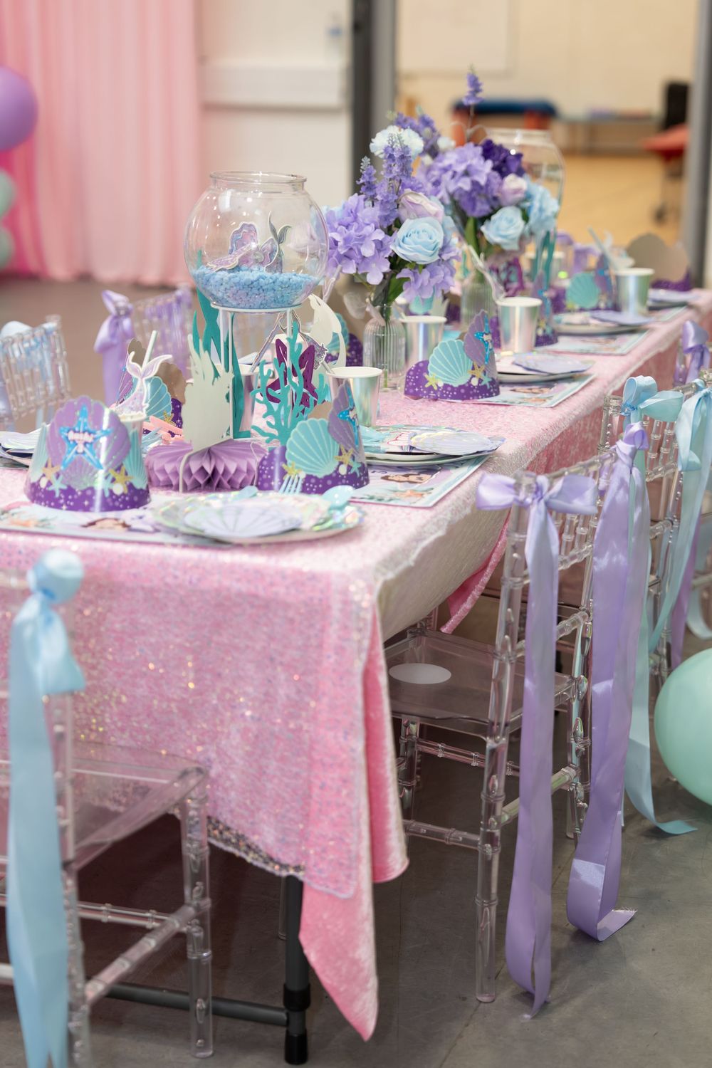 Pink sequin tablecloth mermaid birthday party table with purple flowers, teal accents, and clear acrylic chairs with ribbon bows.