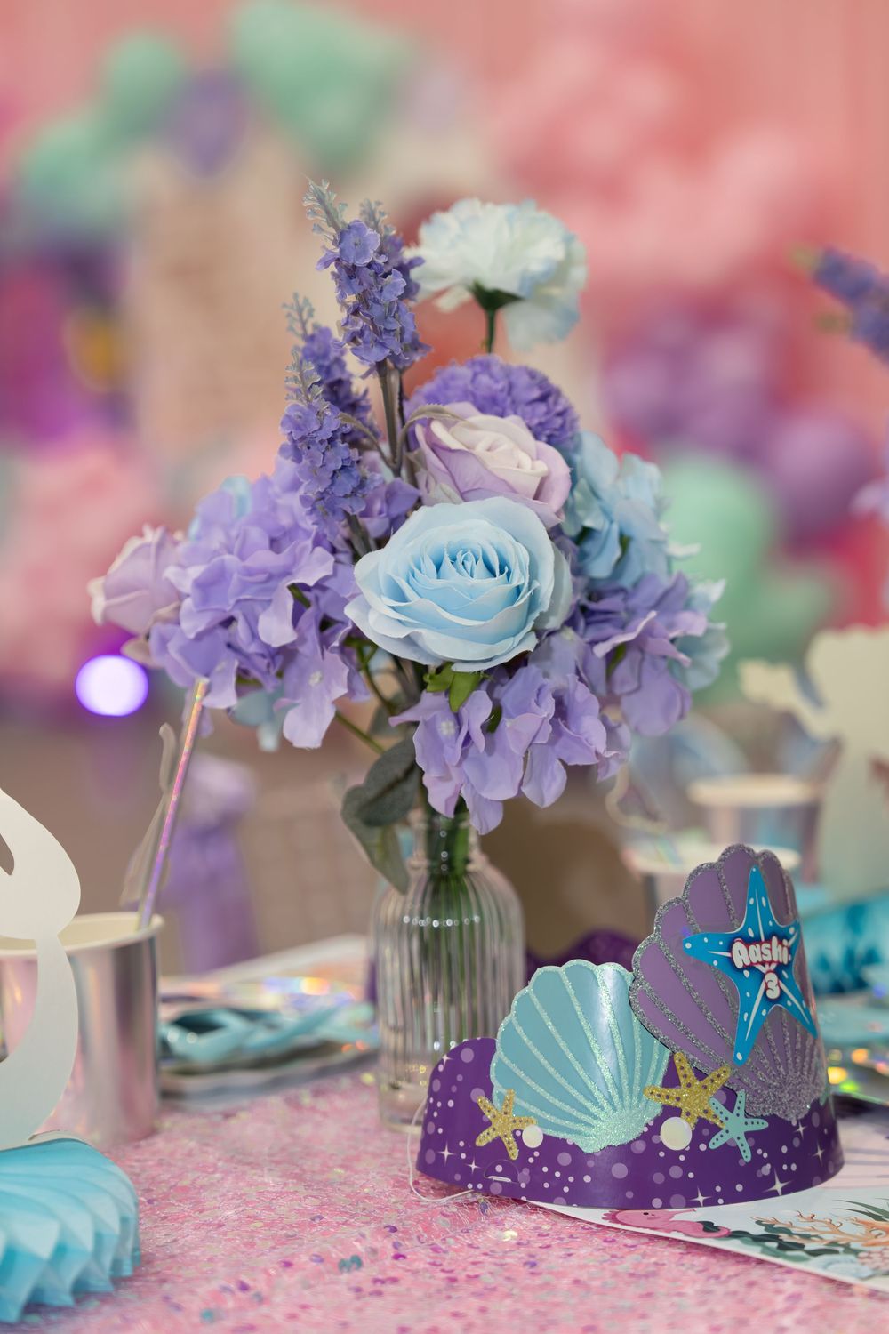 Purple and blue floral centerpiece with mermaid shell crown party decoration on glittery pink table.